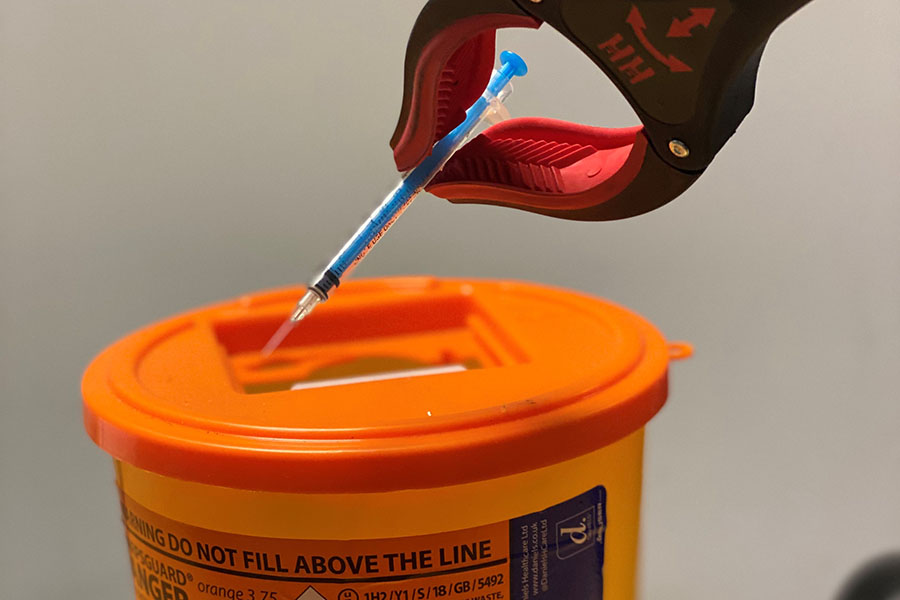 A hypodermic needle being placed into a sharps disposal bin using a picker tool